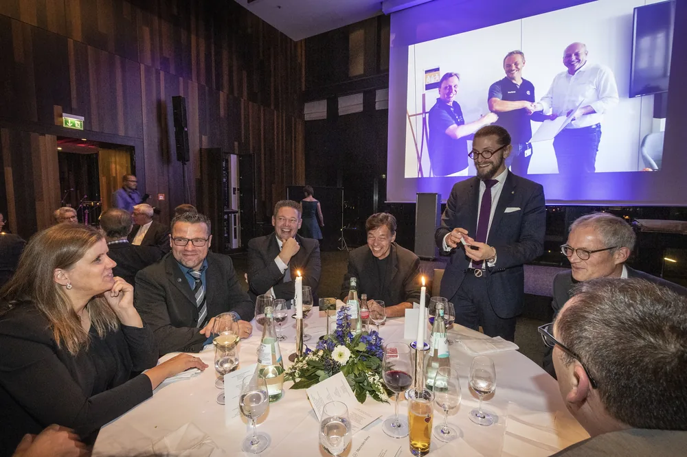 Zauberer Simabu in der Tischrunde eines Gala-Dinners, Leinwand im Hintergrund